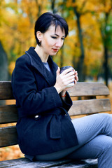 Beautiful woman sitting with coffee and dreaming in autumn city park, fall season, yellow leaves