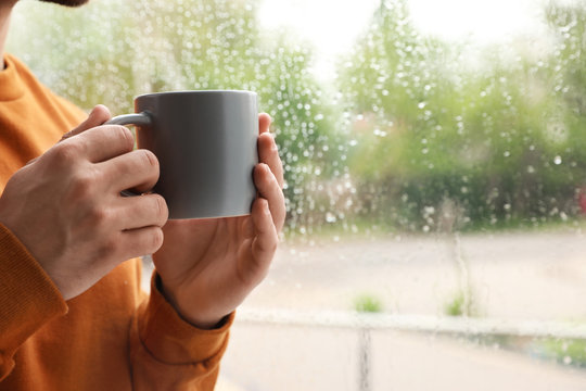 Young man with cup of coffee near window indoors on rainy day, closeup