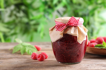 Glass jar of sweet jam with ripe raspberries on wooden table against blurred background. Space for text
