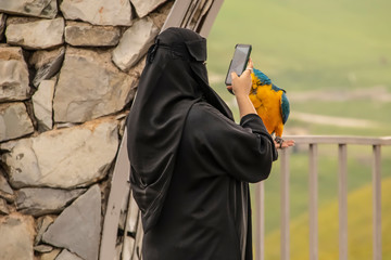Woman in crepe textured burka at scenic outlook taking photo with a smartphone of a parrot she is...