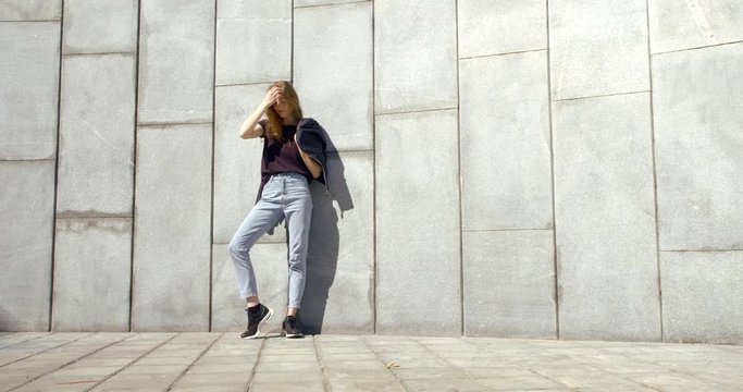 teenage girl is posing near tiled wall in city in sunny day, holding her black jacket