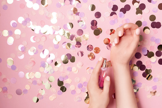 Luxury perfume concept. Female hands holding stylish bottle of perfume on pastel pink background with colorful round paper confetti. Creative trendy flat lay with space for text. Holiday concept.