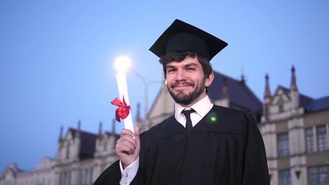 Guy in graduation outfit smiling and showing diploma to camera.
