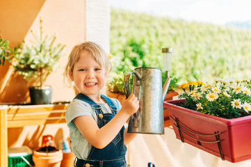 Adorable 3-4 years old kid girl watering yellow daisy flowers on sunny balcony, homework activity...
