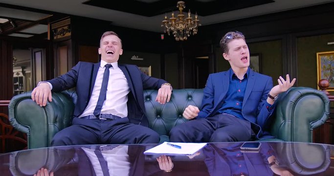 Two young businessmen are sitting in a green leather armchair in luxury apartments in suits and playing imaginary musical instruments. Have fun, funny, music, inspiration, weird, comical
