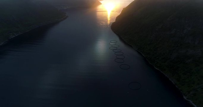 Aerial vertical pan shot of fjord salmon farm and midnight sun under thick clouds in northern Norway island Senja.