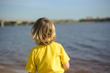 Fototapeta na wymiar Blond kid walking on the beach in light clothes. Warm summer day. A lonely boy on the river Bank. Emotions. Walk with children. rear view. A yellow t-short.