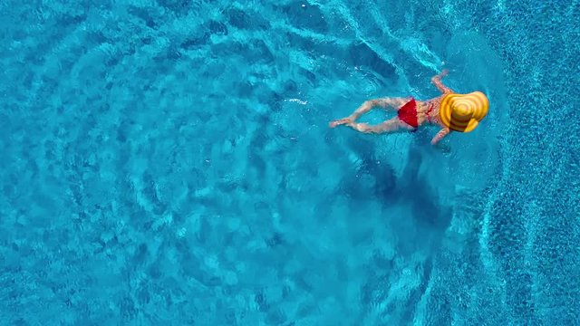 View from the top as a woman in a red swimsuit and a big yellow hat swims in the pool