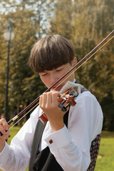 Stylish Young man, a student of music college, plays the violin in the park on a background of yellow-green leaves. Dressed in a white shirt and black vest.