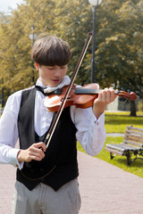 Stylish Young man, a student of music college, plays the violin in the park on a background of yellow-green leaves. Dressed in a white shirt and black vest.