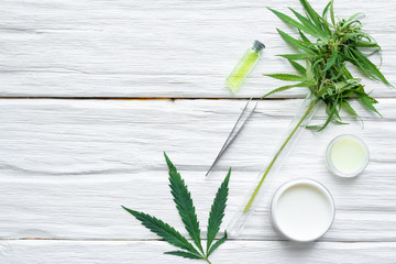 Cannabis cosmetic cream in a jar and a green plant leaf on a white wooden table background with a...