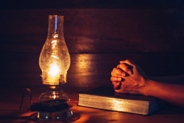 Close up of woman hands praying on bible with oil lamp on wooden table in dark room, Christian...