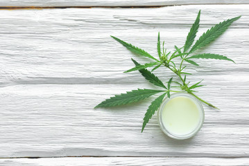 Cannabis cosmetic cream in a jar and a green plant leaf on a white wooden table background. Natural cosmetic.