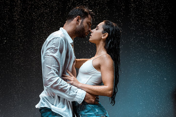 side view of handsome man kissing sexy wet girlfriend under raindrops on black