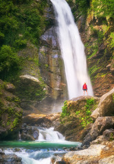 Hiking woman in red jacket stay opposite big waterfall in jungle.