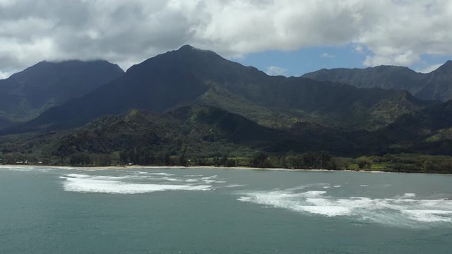 Crystal blue ocean waves with a volcano-like mountain backdrop. Sunny day with clouds hanging above the beach mountains, covered with green terrain. Aerial 4K drone shot panning above the water.