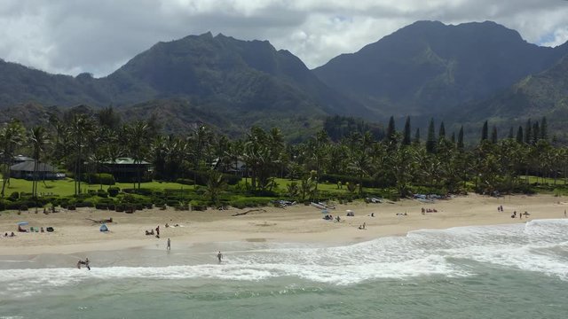 Pacific beach containing crystal blue water, and luscious green mountains in the background. Visitors relax on the beach, next to palm trees, houses, and shops nearby. Aerial orbiting 4K drone shot.