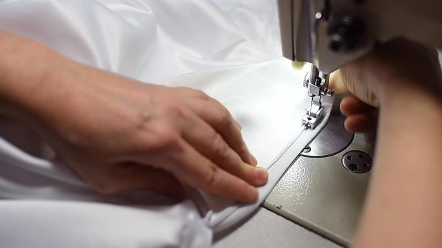 Close up of sewing zipper on white dress. Tailor female hands pulling textile under working mechanism of sewing machine. Built-in hardware lamp turned on. Stitching on accessory parts to garment. POV