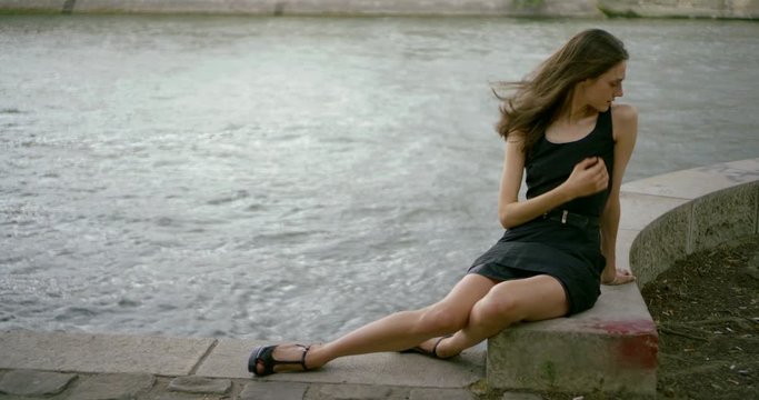 A slender dark-haired girl model in a short black dress sits on a tile near the city river, on her feet she has shoes with heels, the wind blows her hair, she poses.