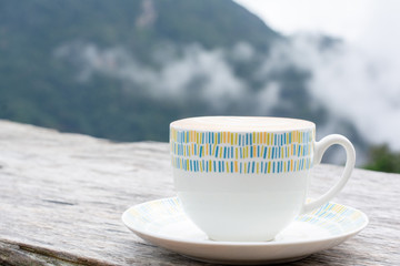 Hot coffee (espresso, cappuccino, latte) in ceramic white cup on wooden table with blurry mountain...