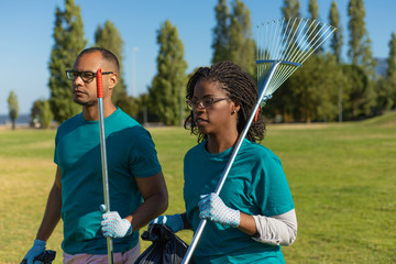 Couple volunteers working in park. Black woman and Latin man walking on grass and carrying rakes...