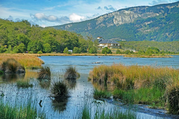 Panoramic view of Tierra del Fuego National Park, showing mountains surrounded by green vegetation and water, against a blue sky.