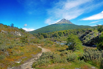 Panoramic view of Tierra del Fuego National Park, showing hills and a volcano surrounded by green vegetation and water, against a blue sky.