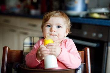 Cute adorable baby girl holding nursing bottle and drinking formula milk. First food for babies. New born child, sitting in chair of domestic kitchen. Healthy babies and bottle-feeding concept