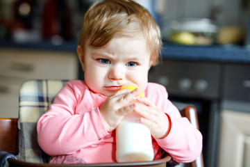 Cute adorable baby girl holding nursing bottle and drinking formula milk. First food for babies. New born child, sitting in chair of domestic kitchen. Healthy babies and bottle-feeding concept