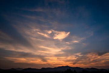 Twilight dramatic blue sky with white clouds and orange sunlight and silhouette of mountains.