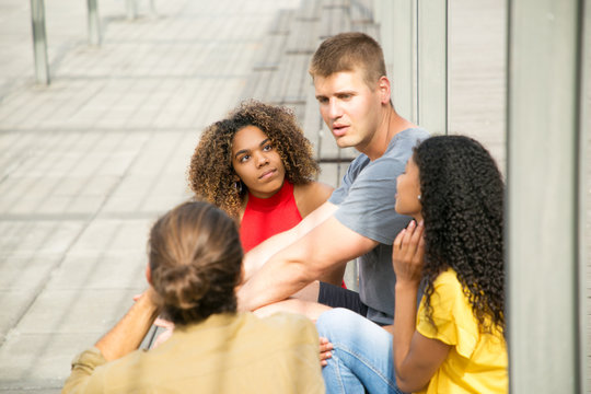 Caucasian guys and mix raced girls meeting outdoors to chat. Interracial group of young people sitting on outdoor staircase, talking, listening, gesturing. Friends meeting concept