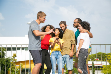 Excited joyful guy telling funny story to his friends outside. Mix raced team of people standing outdoors, leaning on railing, talking, listening, laughing. Friends outdoors concept