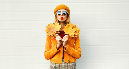 Autumn portrait woman holding yellow maple leaves blowing red lips sending sweet air kiss in french beret posing over gray wall background