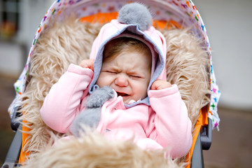 Sad crying little beautiful baby girl sitting in the pram or stroller on autumn day. Unhappy tired...