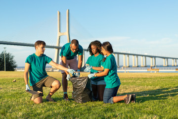 Multiethnic volunteer team removing garbage from grass into plastic bag. Men and women wearing...