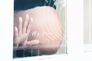 Pregnancy, motherhood, people and expectation concept. Cropped of hand pregnant woman standing by the window with text My love. Copy space for your text.