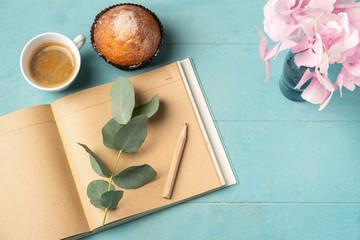 Flat lay, top view of feminine desk. Coffee cup for breakfast, cupcake, empty notebook, branch with eucalyptus leaves and and pink hydrangea. Business planning breakfast concept.
