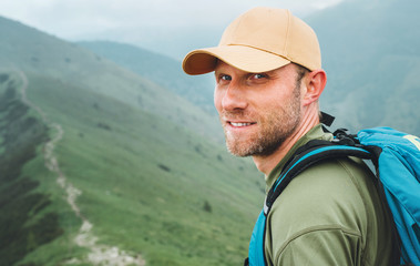 Portrait of backpacker man in baseball cap walking by the foggy cloudy weather mountain range path with backpack. Active sports backpacking healthy lifestyle concept. - obrazy, fototapety, plakaty