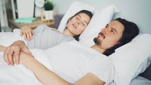 Unhappy man husband is covering head with pillow while wife is snoring in bed disturbing his sleep in bedroom. People, relationship and bedtime concept.