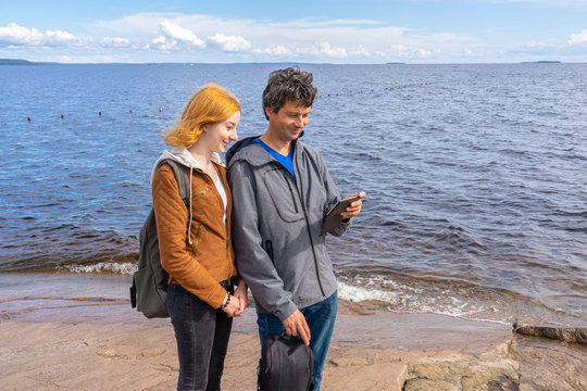 Middle aged man and young lady standing on northern lake shore in summer day. Tourists taking pictures on smartphone. Travelling and discovering distant places of Earth. Onega lake, Karelia, Russia