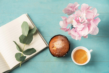 Flat lay, top view of feminine desk. Coffee cup for breakfast, cupcake, empty notebook, branch with eucalyptus leaves and and pink hydrangea. Business planning breakfast concept.
