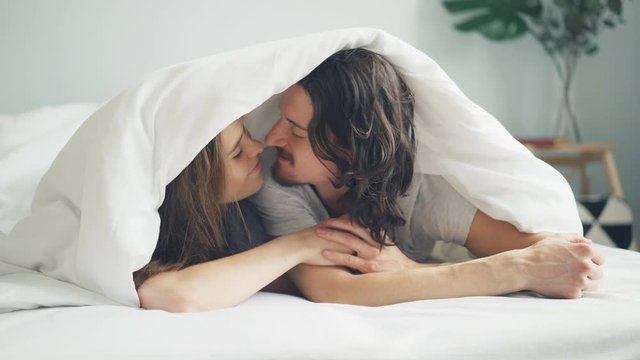 Happy couple man and woman are kissing and rubbing noses under blanket lying on bed at home together expressing love and tenderness. People and affection concept.