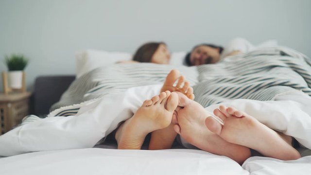 Man and woman couple are lying in bed touching feet talking enjoying bedtime together in cosy bedroom. Relationship, relaxation and apartment concept.