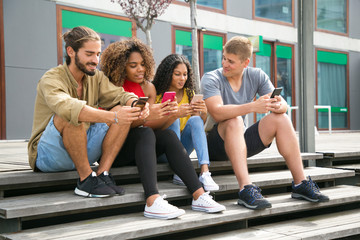 Fototapeta na wymiar Happy friends meeting outside and using their phones. Interracial group of people sitting outdoors and using smartphones. Communication concept