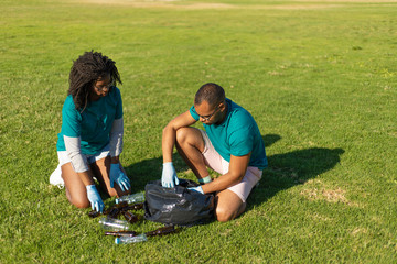 Outdoor cleaners collecting empty bottles into plastic bag. Black woman and Latin man wearing...