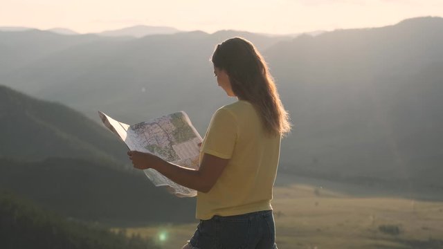 Camera movement around a tourist girl who stands on the edge of a cliff and holds a map in her hands