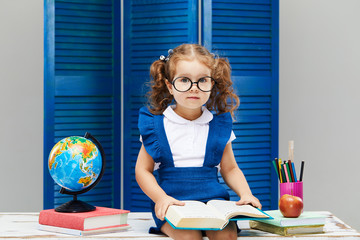 Smart toddler kid. Back to school and happy time! Cute industrious child in glasses is sitting at a desk indoors. Kid is learning in class on background of blue wall. Girl reading the book.