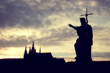 Naklejka premium Silhouette of St. John the Baptist statue on Charles Bridge in Prague with St. Vitus Cathedral in background at sunset.