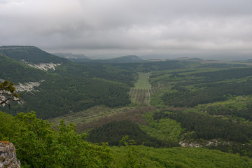 view of the valley of green with trees and grass, on a cloudy day with clouds in the sky, and mountains with flat peaks of white and gray.