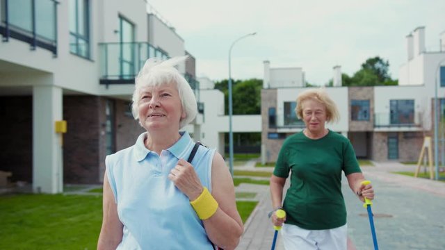 Pretty Mature Woman In Sportswar Walking In The Street. Her Old Sportive Female Friend Walking Fast With Ski Poles In Background. Friendship Has No Age.